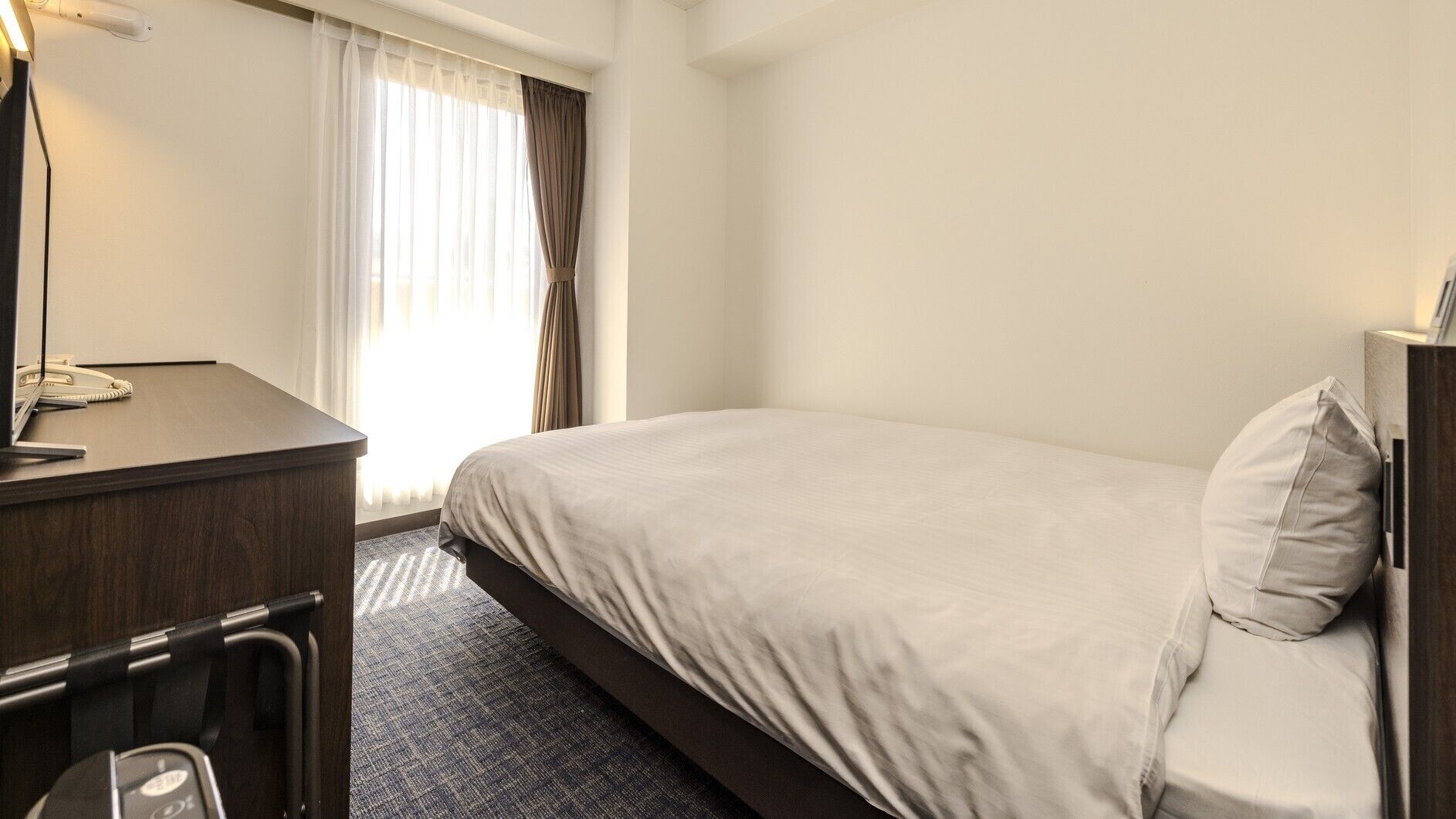 A hotel room with a bed, a desk with a telephone, and a window with curtains.