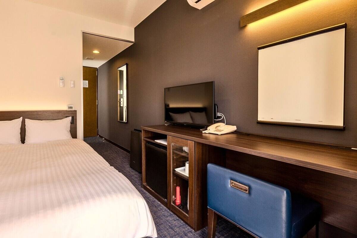 A hotel room with a bed, a desk with a telephone, a mirror, and a wall-mounted TV.