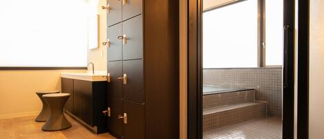 A modern bathroom with a wooden floor, a dark cabinet with multiple drawers, a small sink, and a view of a tiled shower area through an open door.