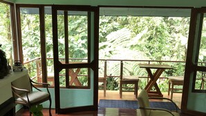 A room with wooden flooring, a sofa, a dining table, and chairs. Large windows offer a view of greenery outside. - Agua Inn (Arenal)