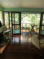Habitación con suelo de madera, un sofá, una mesa de comedor y sillas. Amplias ventanas ofrecen vistas a la vegetación exterior.