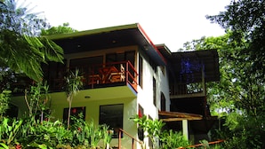 A two-story building with a balcony, surrounded by lush greenery and a stone wall. - Agua Inn (Arenal)