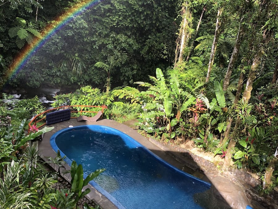 Une piscine entourée d’une végétation luxuriante, avec un arc-en-ciel bien visible.