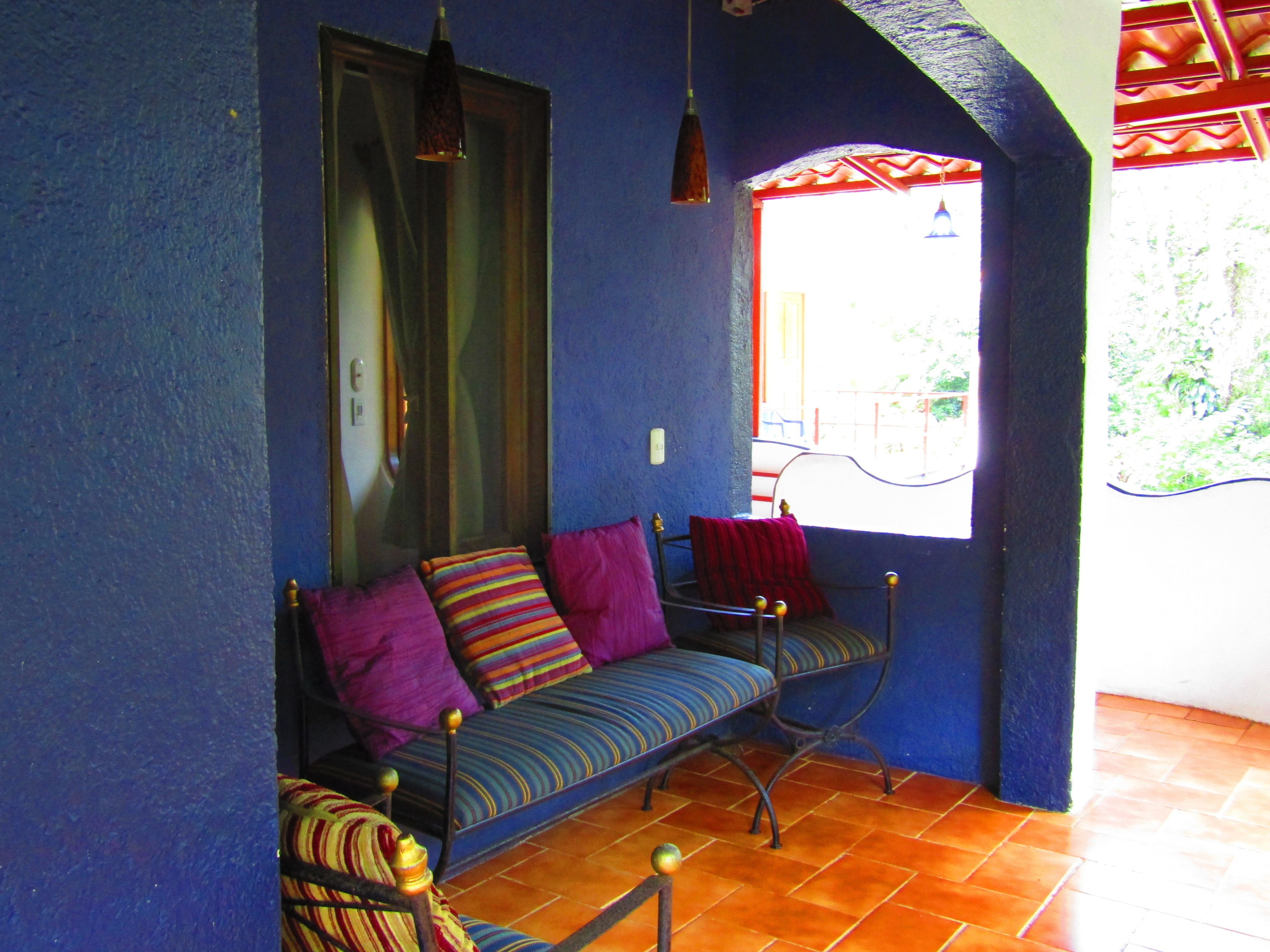 A room with a blue wall, a window with a view, a sofa with colorful cushions, and a hanging light fixture.