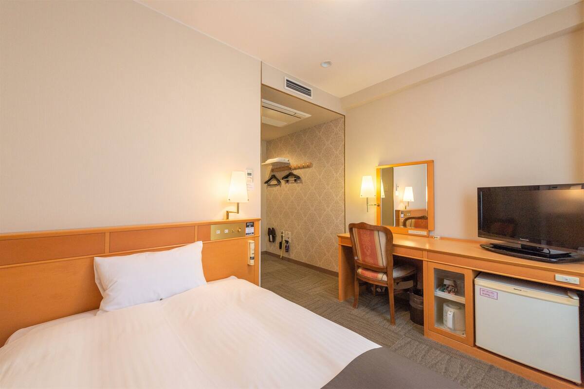A hotel room with a bed, a desk with a television, a chair, a mini-fridge, and a mirror.