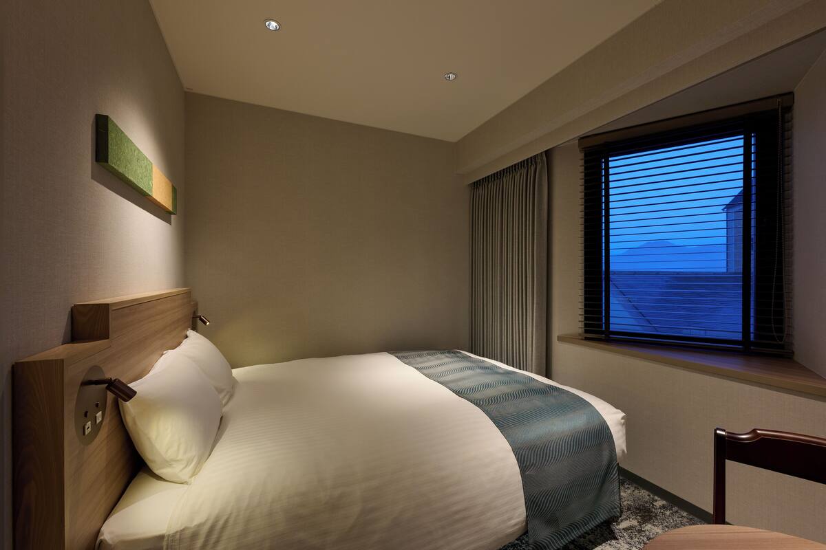 A hotel room with a bed, a chair, a window with blinds, and a ceiling light.