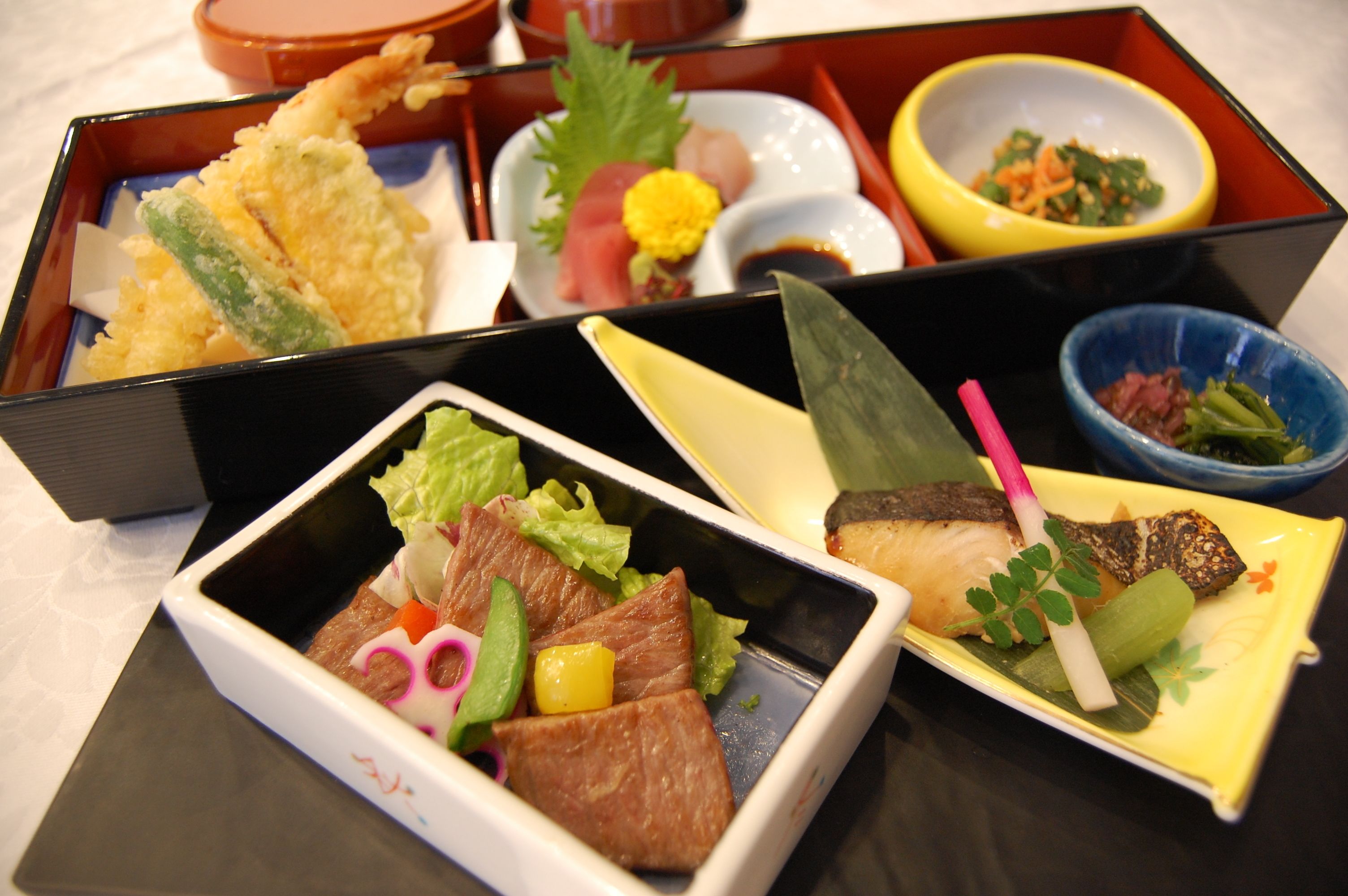 A variety of Japanese dishes in separate compartments of a bento box, including sushi, sashimi, and tempura.