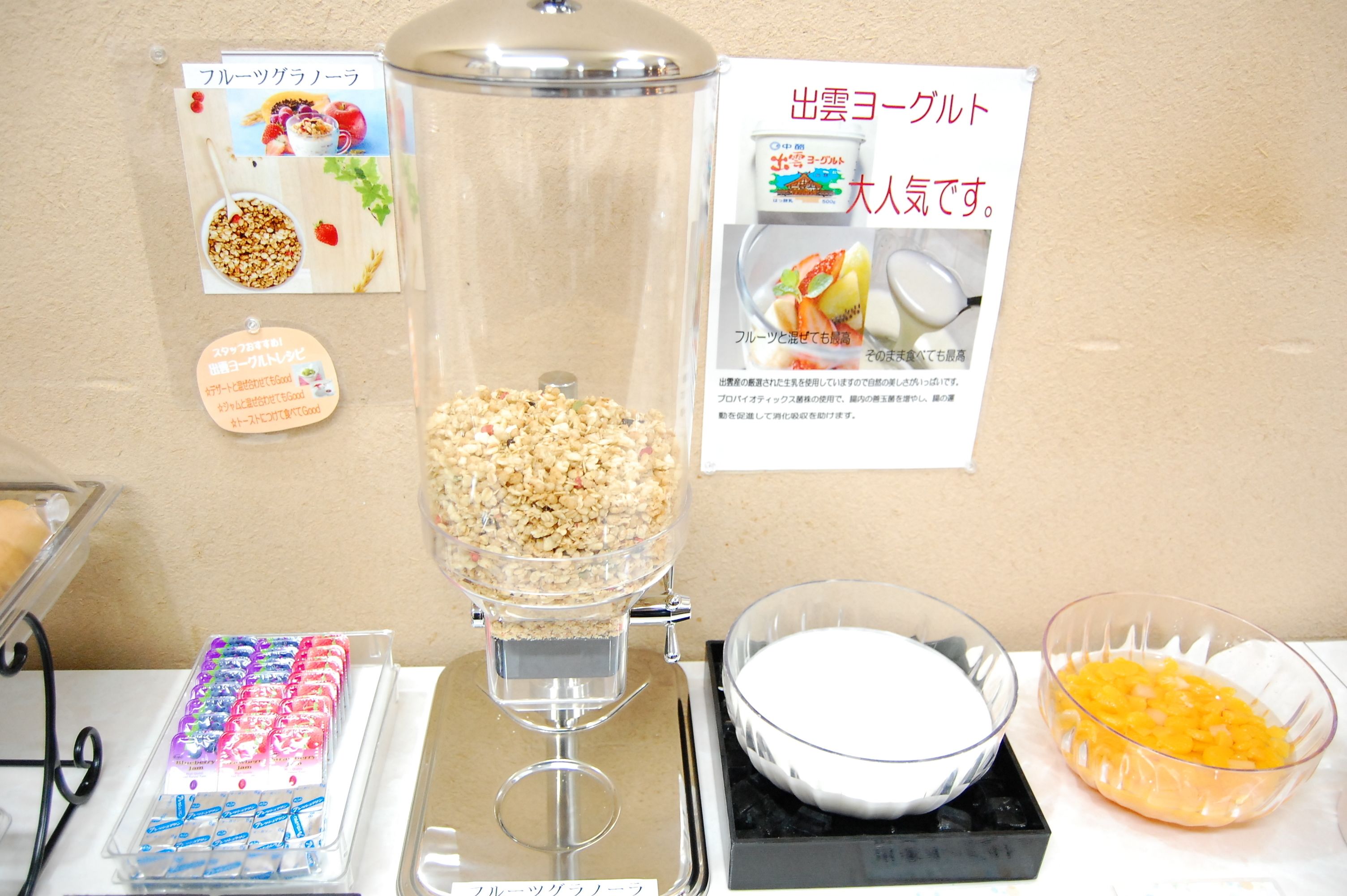 A food station with a bowl of cereal, a bowl of yogurt, and a container of granola.