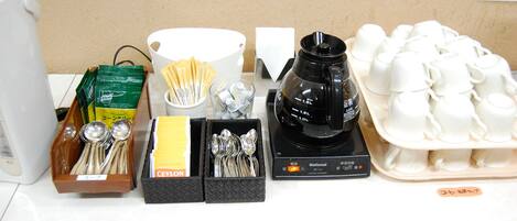 A coffee station with a coffee maker, cups, spoons, and a tray of white cups.