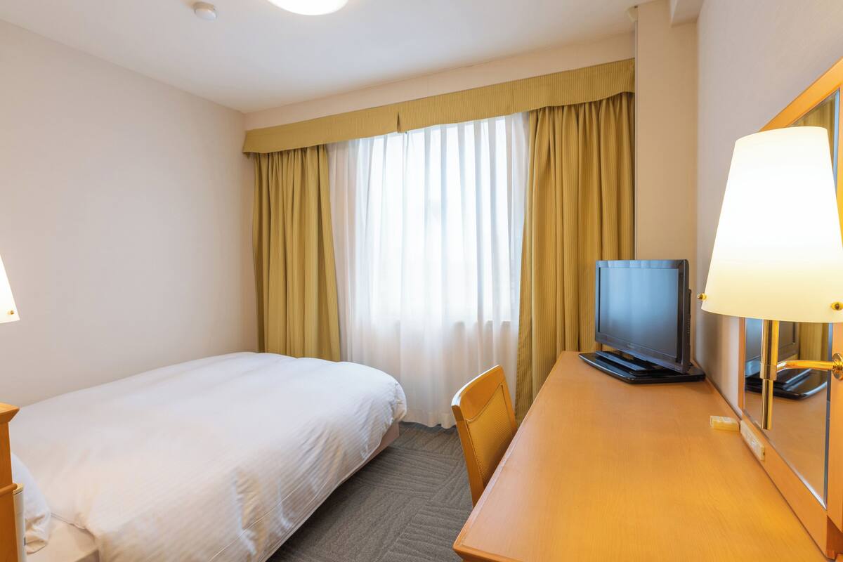 A hotel room with a single bed, a desk with a television, and a lamp.
