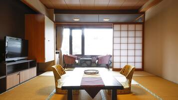 Una habitación japonesa tradicional con suelo de tatami, una mesa baja cubierta con un felpudo de tatami, dos sillas, un televisor sobre un mueble y un panel corredizo shoji.