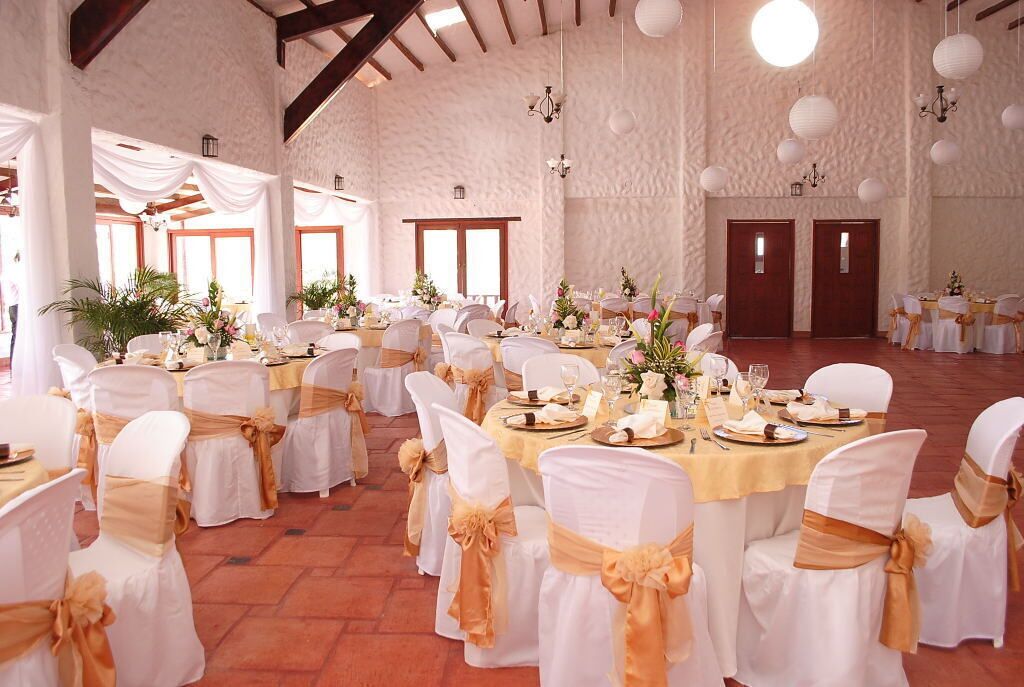 Un salón de banquetes con mesas redondas, sillas blancas adornadas con cintas doradas y paredes blancas.