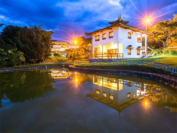 A traditional building with a reflection in a calm water body, surrounded by greenery and illuminated by ambient lighting.