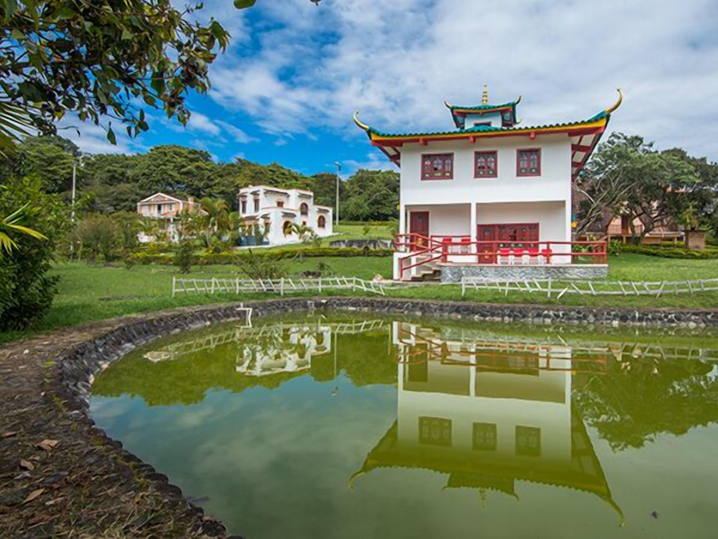 A traditional building with a reflection in a pond, surrounded by greenery and other structures.