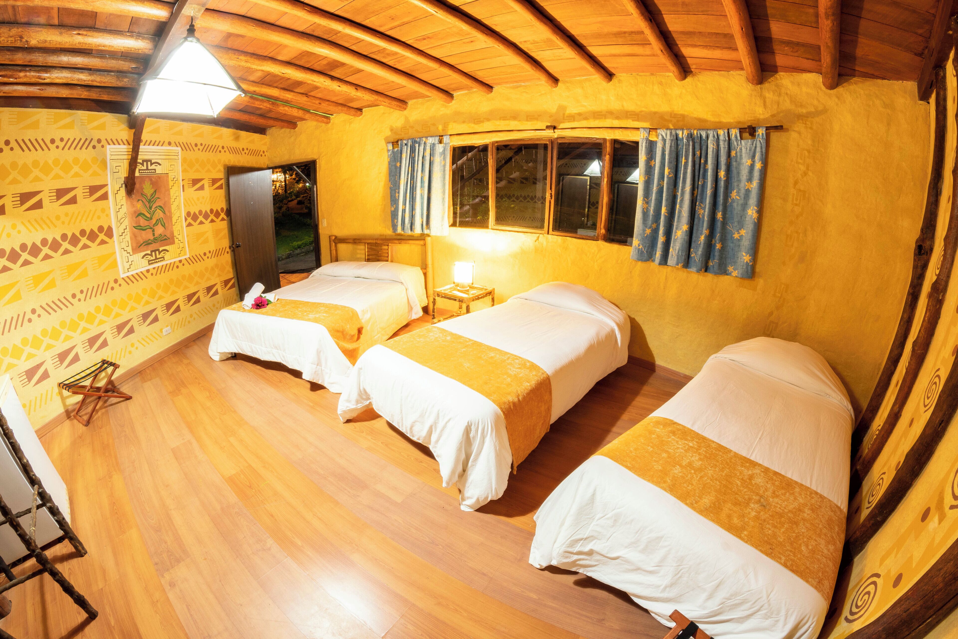 Una habitación con dos camas, piso de madera, techo de madera, una ventana con cortinas y una pared con papel tapiz estampado.