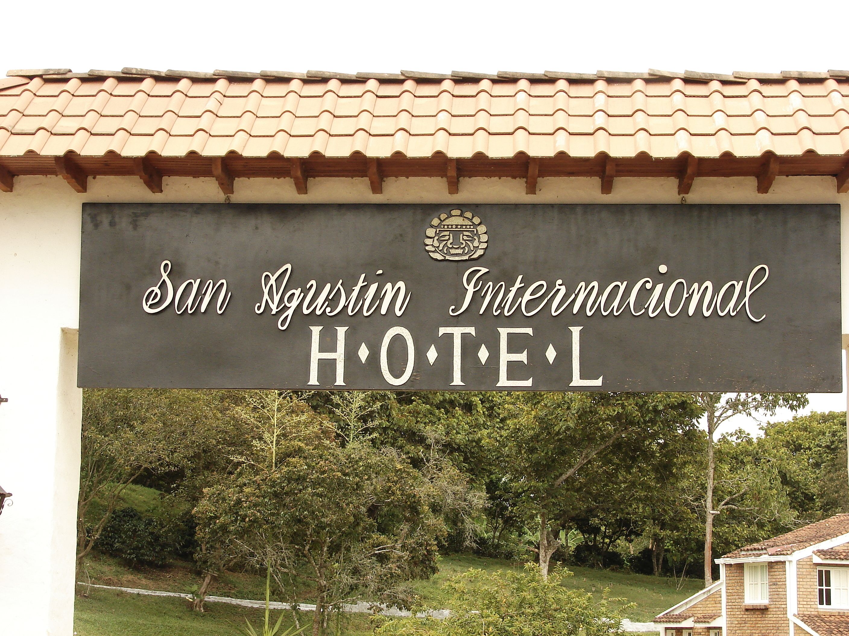 A sign for San Agustin Internacional Hotel with a tiled roof and greenery in the background.