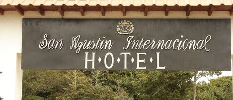 Señal para el Hotel San Agustin Internacional, con techo de tejas y follaje al fondo.