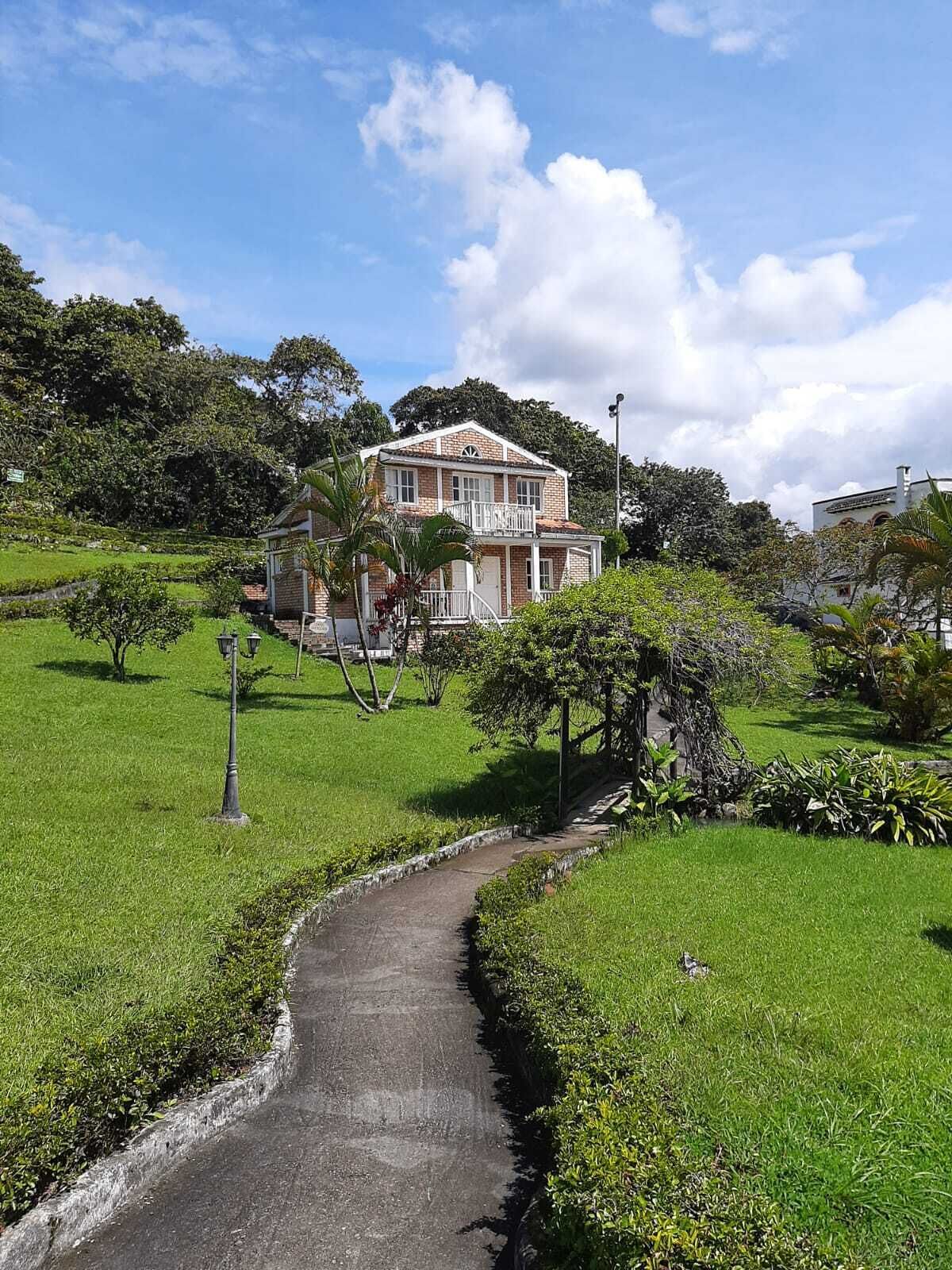 Una casa de dos pisos con un balcón, rodeada de abundante vegetación y un camino empedrado.