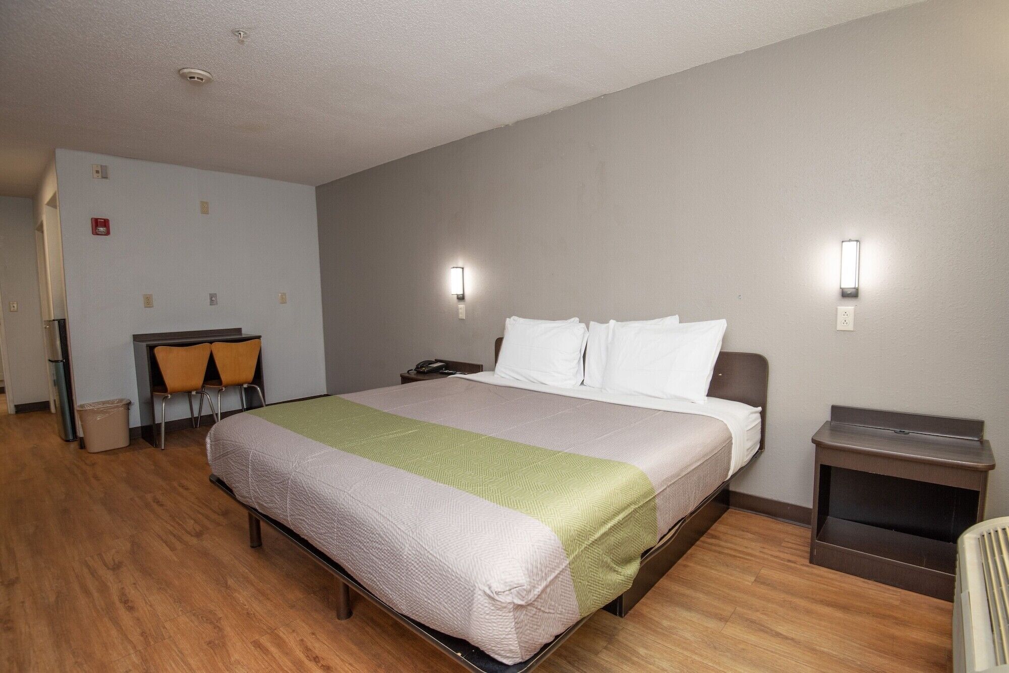 A hotel room with a bed, a desk with two chairs, a trash bin, and a nightstand.