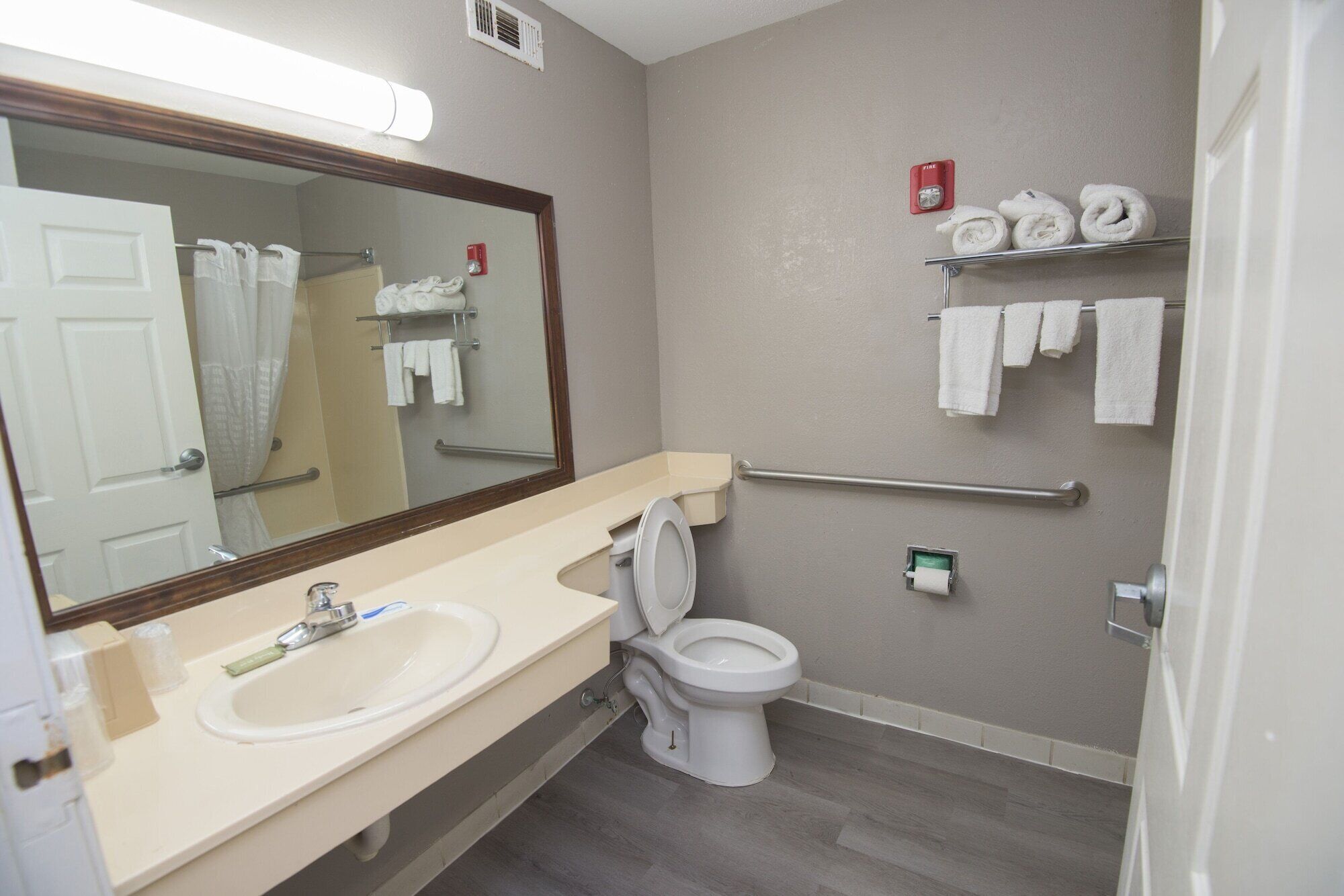 A bathroom with a sink, toilet, and a mirror. There are towels on a rack and a fire alarm on the wall.