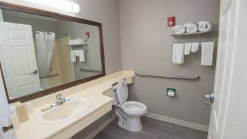 A bathroom with a sink, toilet, and a mirror. There are towels on a rack and a fire alarm on the wall.