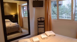 Hotellihuone, jossa on sänky, peili, televisio, ikkuna verhoilla ja pyyheteline pyyhkeineen.