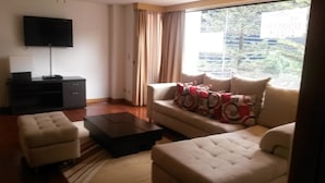 Una sala de estar con sofá, reposapiés, mesa de centro, televisor y una ventana con cortinas.