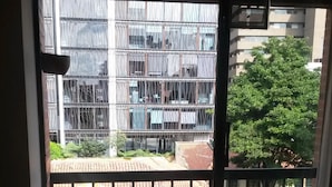 Vista desde una ventana con baranda metálica, con vista a un edificio de varios pisos y un árbol.
