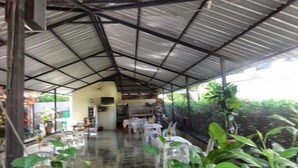 Un área de comedor al aire libre con techado, mesas y sillas, una escalera que sube a un nivel superior y plantas en macetas.