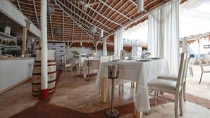 A restaurant with white tables and chairs, a bar area, and a thatched roof. - Corales Colonial Apartament With Private Beach (Punta Cana)