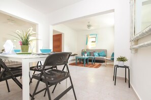 Un coin repas avec une table blanche, des chaises noires et une plante en pot. On aperçoit un canapé avec des coussins de couleur turquoise à l’arrière-plan.