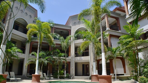 A multi-story hotel with a central courtyard featuring a swimming pool, surrounded by palm trees and lounge chairs. - Macaws Jaco Condos No.12 (Jaco)