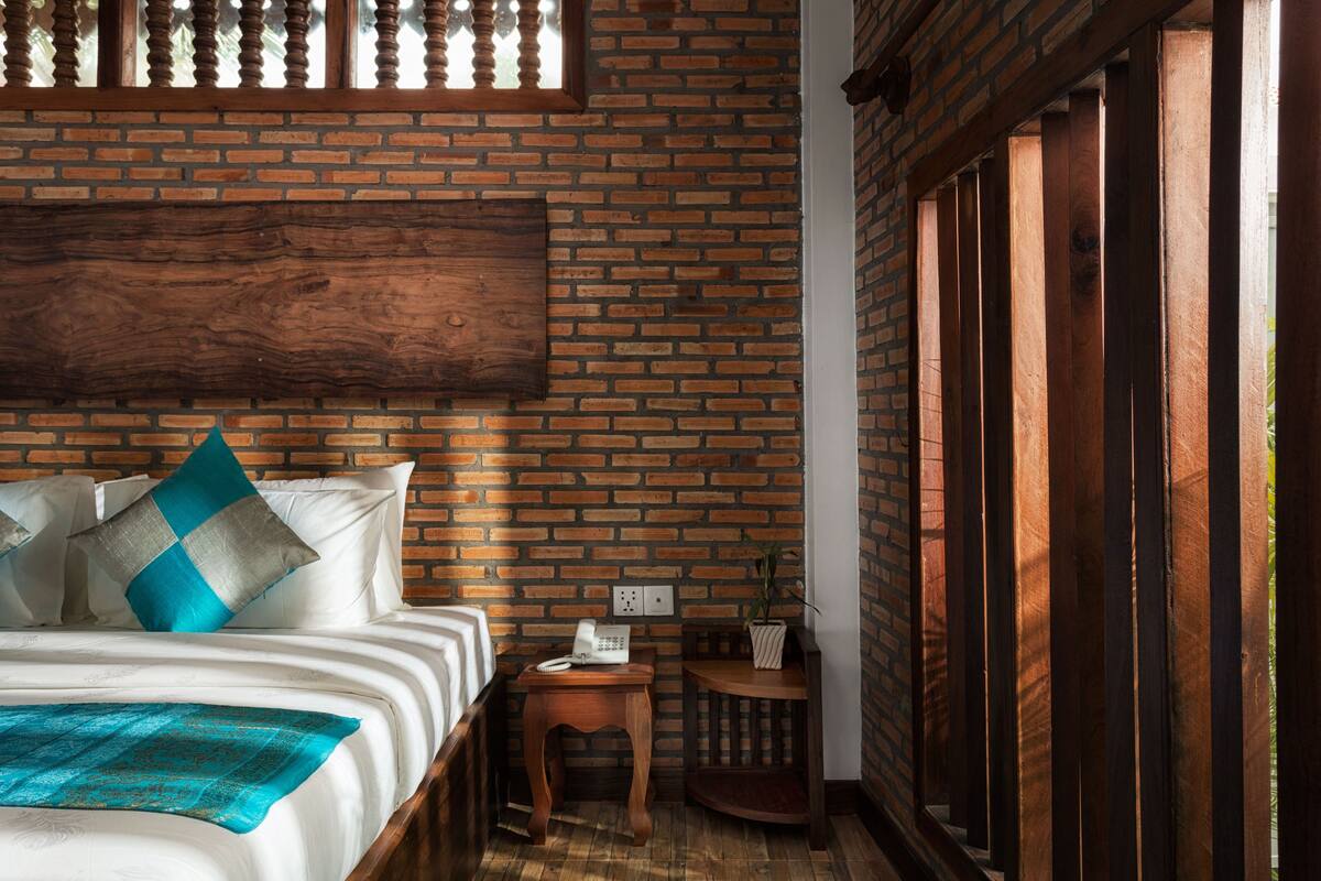 Ein Schlafzimmer mit Backsteinwand, Holzfußendecke, einem Bett mit weißen Leinen, blauen und grauen Kissen, einem Nachttisch mit Telefon und einem kleinen Holzschemel.