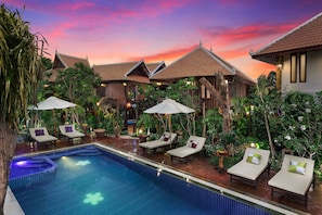 Un área de piscina con sillones de descanso y sombrillas, rodeada de exuberante vegetación y arquitectura tradicional.