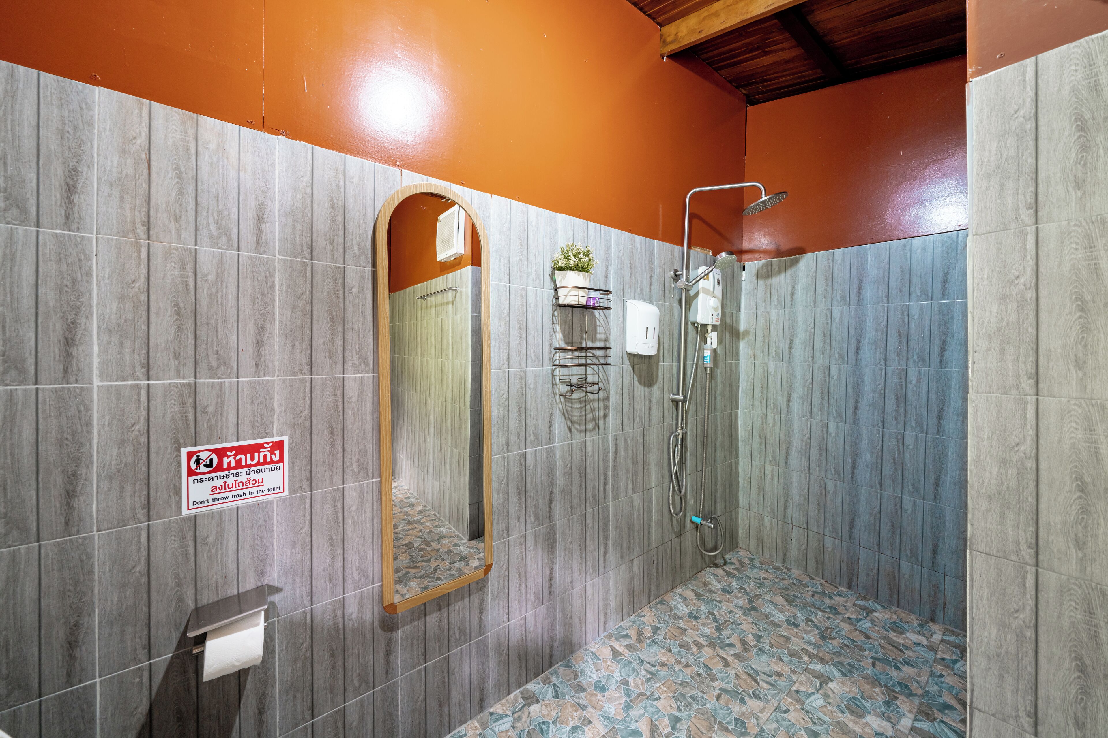 Un baño con paredes grises y anaranjadas, suelo de baldosas, ducha, inodoro y espejo.