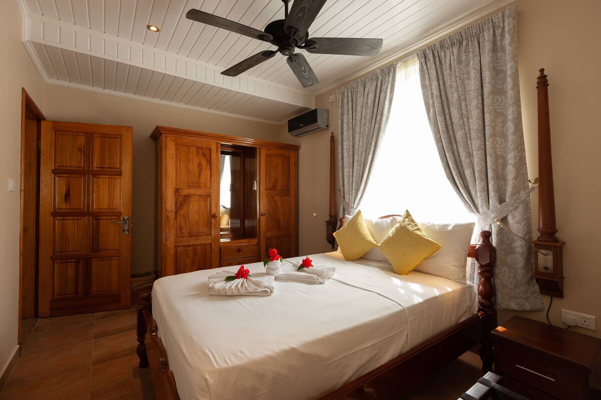A hotel room with a large bed, wooden wardrobe, ceiling fan, and a window with curtains.