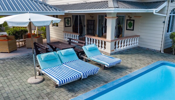 Un espace aménagé au bord de la piscine avec des chaises longues et une table, une terrasse couverte avec des chaises et une table, et une maison dotée d’un balcon.