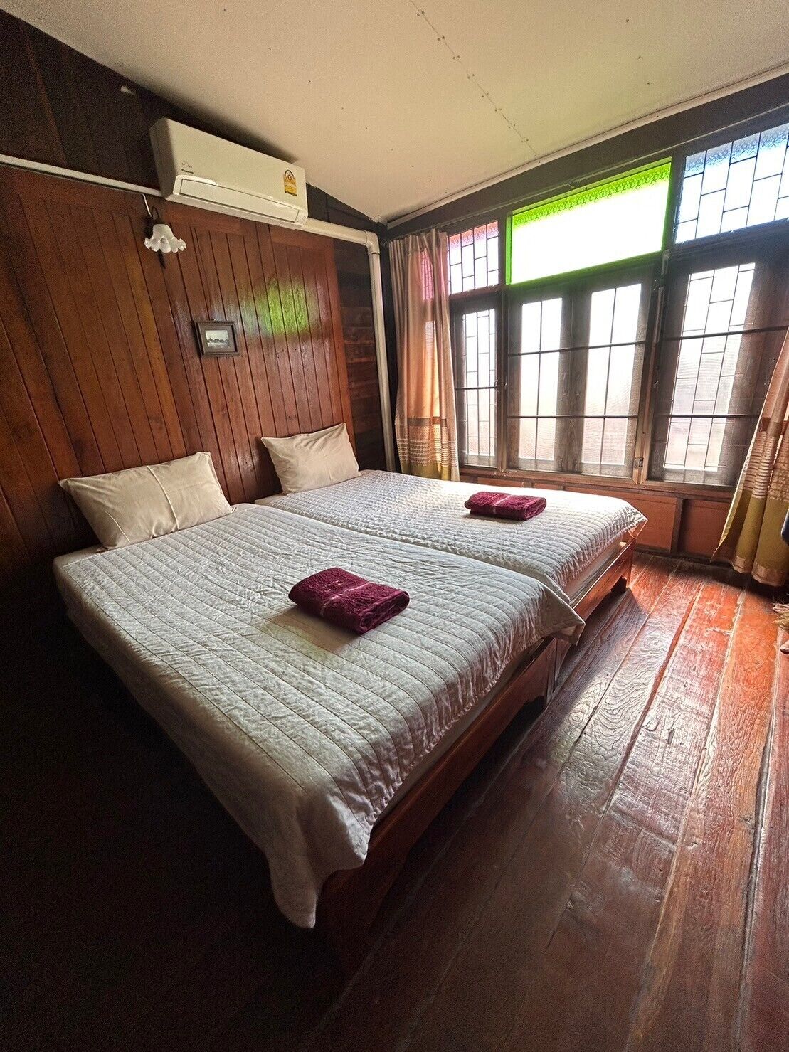 A room with two beds, wooden walls, a window with curtains, and an air conditioning unit.