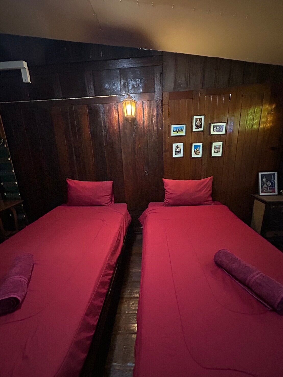 A room with two beds, wooden walls, a wall-mounted light, and framed pictures.