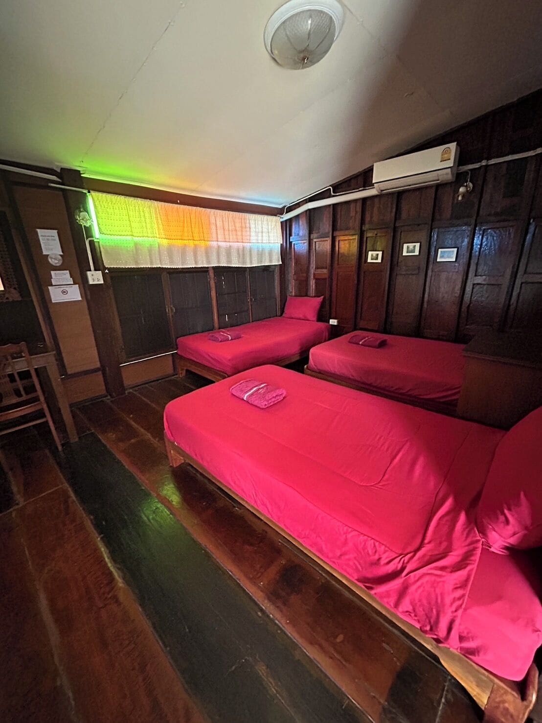 A room with two beds, wooden walls, a ceiling fan, and a window with a colorful curtain.