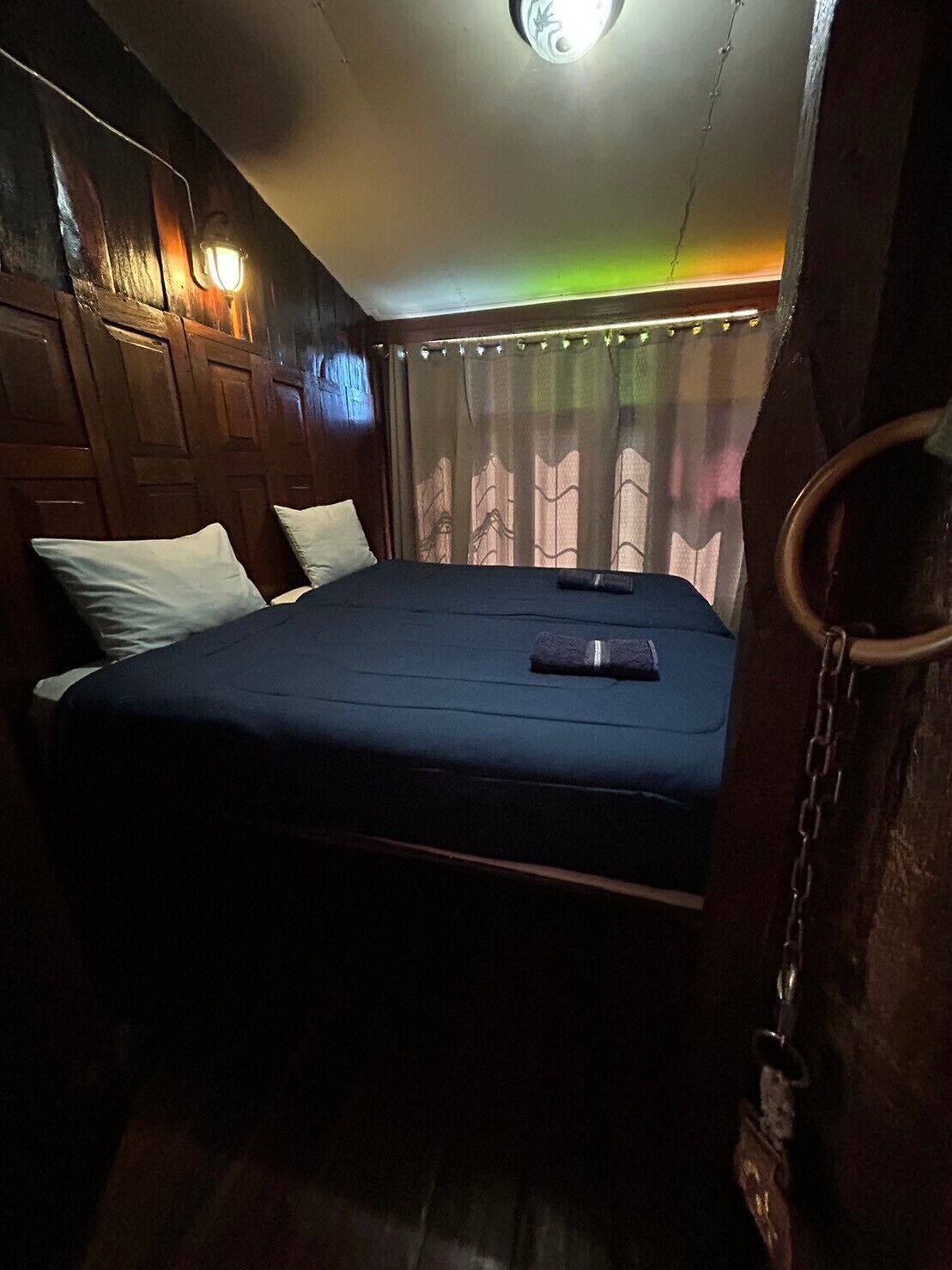A single bed with a dark blue mattress, white pillows, and a black object on the bed. The room has wooden paneling on the walls and a curtain with a pattern.