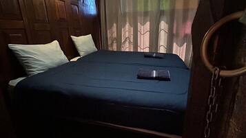 A single bed with a dark blue mattress, white pillows, and a black object on the bed. The room has wooden paneling on the walls and a curtain with a pattern.