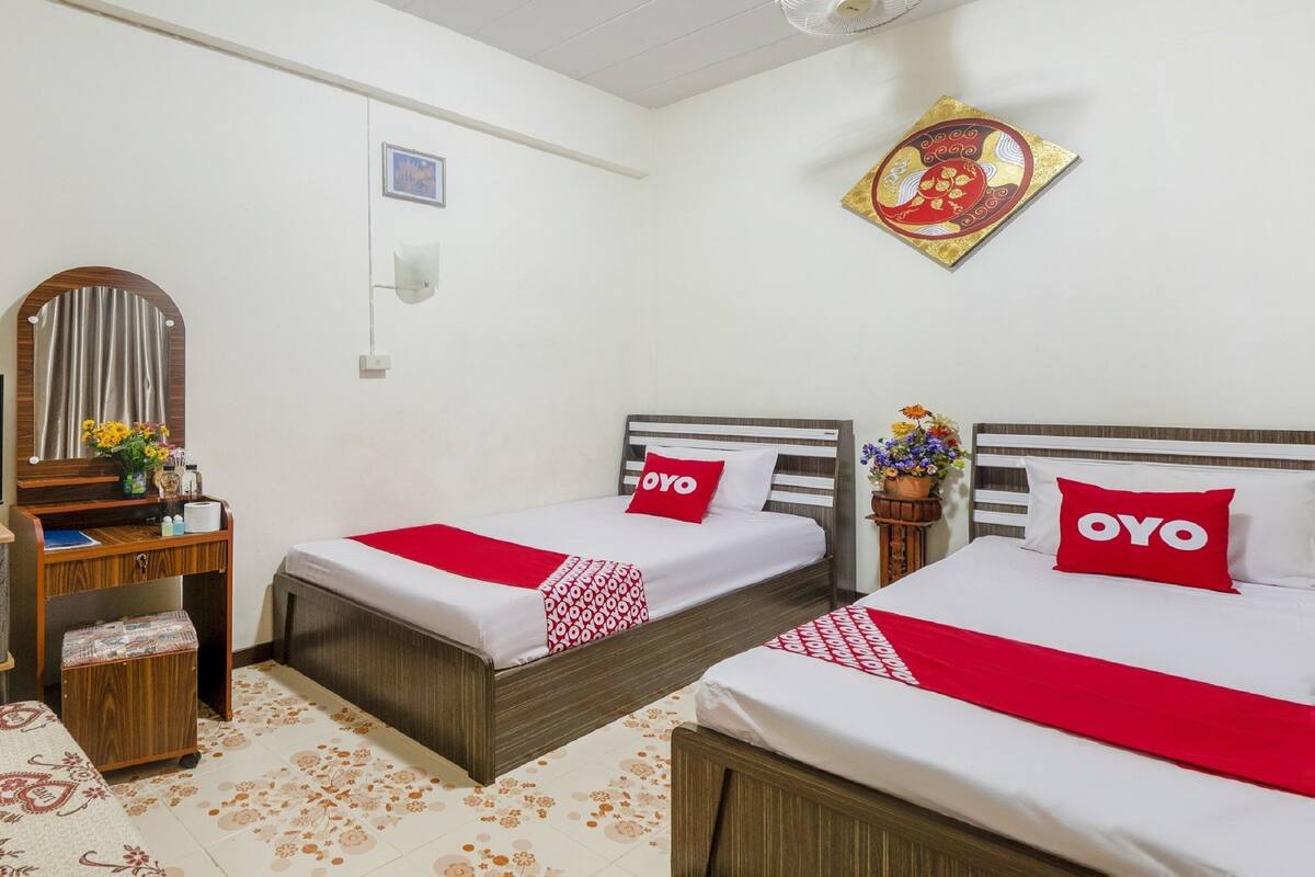 Une chambre d’hôtel avec deux lits, une commode en bois, un miroir, un ventilateur fixé au mur et un élément décoratif mural.