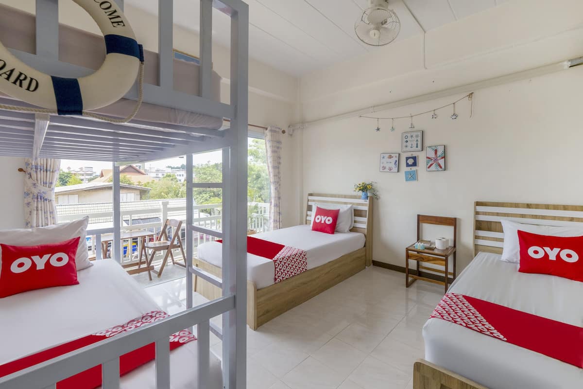 Une chambre avec deux lits, chacun recouvert d’une courtepointe rouge et blanche et agrémenté d’oreillers portant l’inscription « OYO ». On y trouve une chaise en bois et une petite table sur laquelle sont posés une plante et une bouteille. Les murs de la chambre sont blancs et elle dispose d’un balcon offrant une vue sur les bâtiments et les arbres.