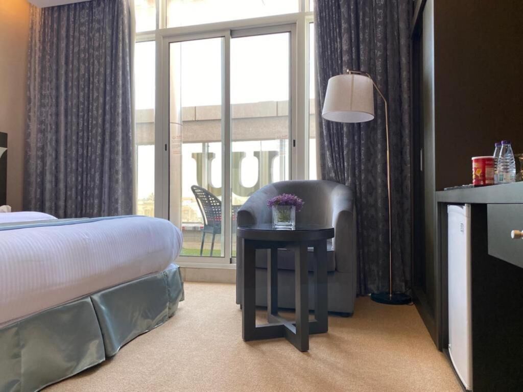 Habitación de hotel con cama, una mesita de noche con un florero, una silla, un escritorio y una ventana con cortinas.