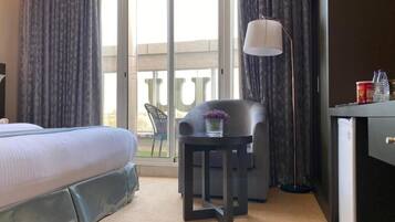 Habitación de hotel con cama, una mesita de noche con un florero, una silla, un escritorio y una ventana con cortinas.