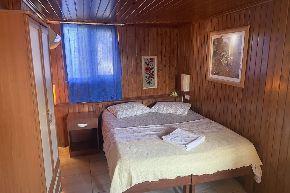 Un dormitorio con paneles de madera, una cama, una mesita de noche, una lámpara y una ventana con cortinas azules.