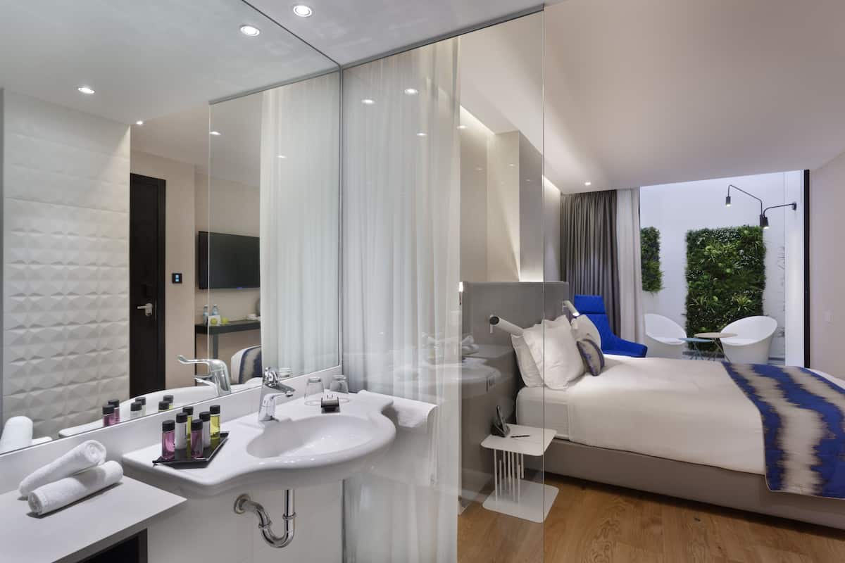 Ein modernes Hotelzimmer mit einem großen Spiegel, einem weißen Waschbecken, einem Bett mit blauen Akzenten und einem kleinen Tisch mit einer Lampe.