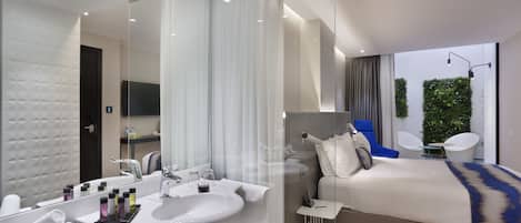 A modern hotel room with a large mirror, a white sink, a bed with a blue accent, and a small table with a lamp.