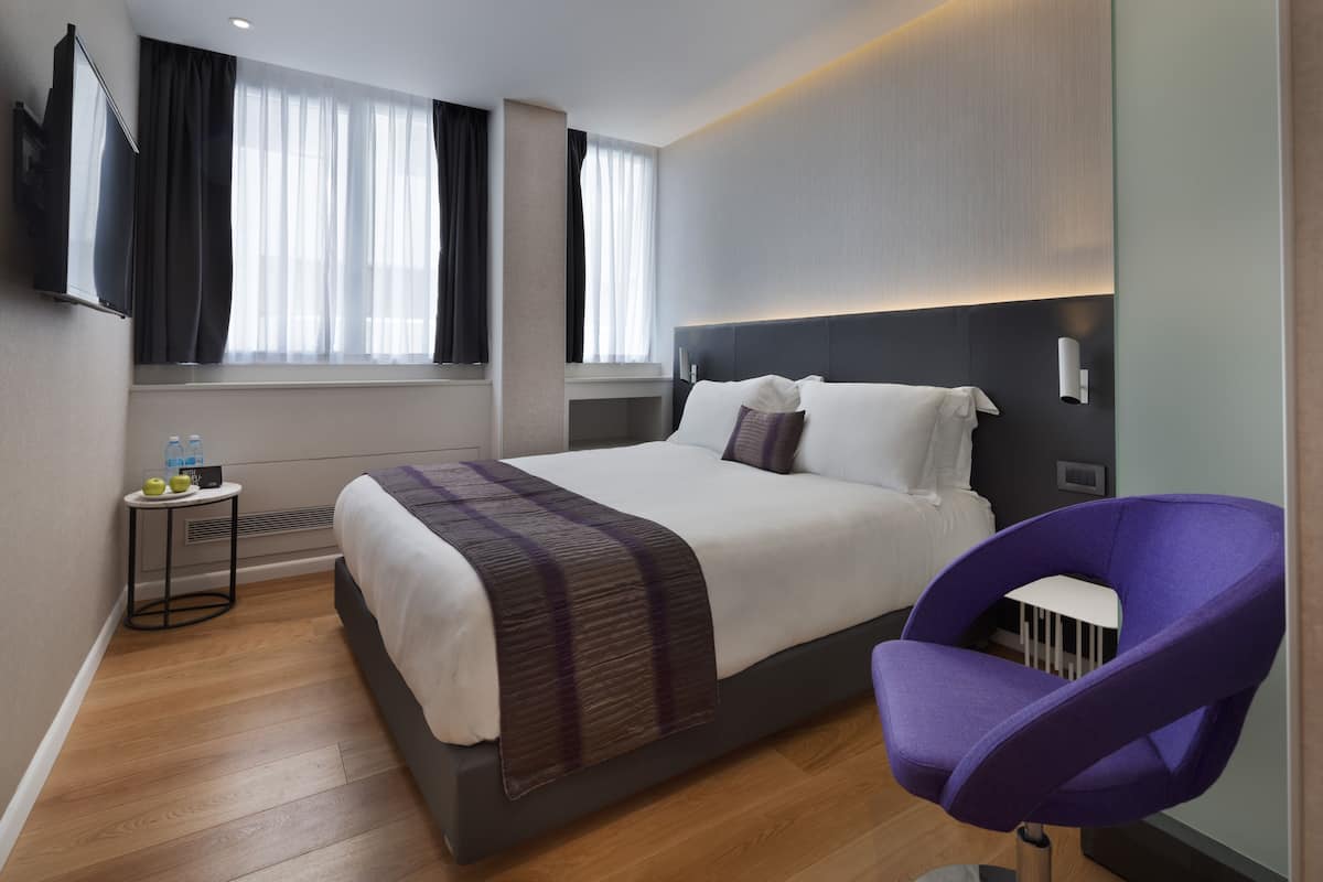 Ein modernes Hotelzimmer mit einem großen Bett, einem Nachttisch mit Flasche und Glas sowie einem violetten Sessel.